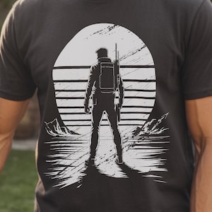 Puede incluir: Camiseta negra con un gráfico de una persona frente a una puesta de sol y montañas. La persona lleva una mochila y un rifle. La palabra "BLACK" está impresa en la parte inferior de la camiseta.