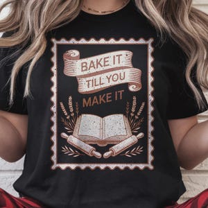 May include: Black t-shirt with a rectangular graphic featuring the text "BAKE IT TILL YOU MAKE IT" above an open cookbook and rolling pins. The design is in shades of brown and white. The shirt is worn with red and black plaid pajama pants.
