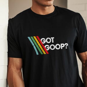 Puede incluir: Camiseta negra con el texto blanco "GOT GOOP?", acompañada de un diseño retro de arcoíris. La camiseta es de cuello redondo.