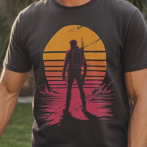 Puede incluir: Camiseta negra de cuello redondo con un gráfico de una persona frente a una puesta de sol y una cordillera. La puesta de sol es naranja y amarilla con rayas rojas y rosas. La palabra "BLACK" está impresa en blanco.