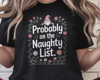 Peut inclure: T-shirt noir avec l'inscription "Probably on the Naughty List". Le motif comprend un gnome avec un chapeau rose, entouré de fleurs et de feuilles. Le bas du t-shirt affiche le mot "BLACK".