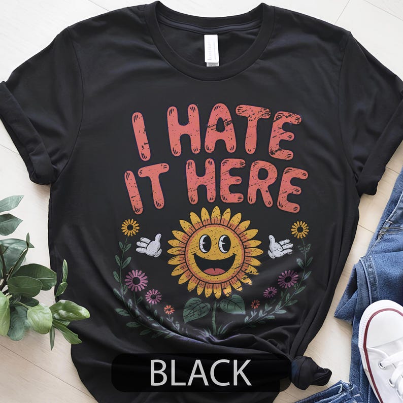 May include: Black t-shirt with the text "I HATE IT HERE" in coral letters above a smiling sunflower graphic. The shirt also features the word "BLACK" at the bottom.