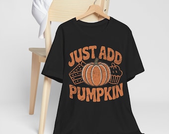 May include: Black t-shirt with the phrase "JUST ADD PUMPKIN" in orange, along with a pumpkin, pie, and cupcake graphic. The shirt is hanging on a wooden hanger.