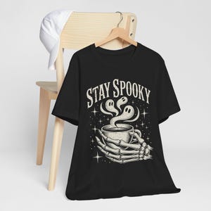 May include: Black t-shirt with a Halloween graphic. The design shows a skeleton hand holding a coffee cup with ghost-shaped steam, and the words "Stay Spooky" in a vintage font. A white baseball cap hangs on a wooden chair.