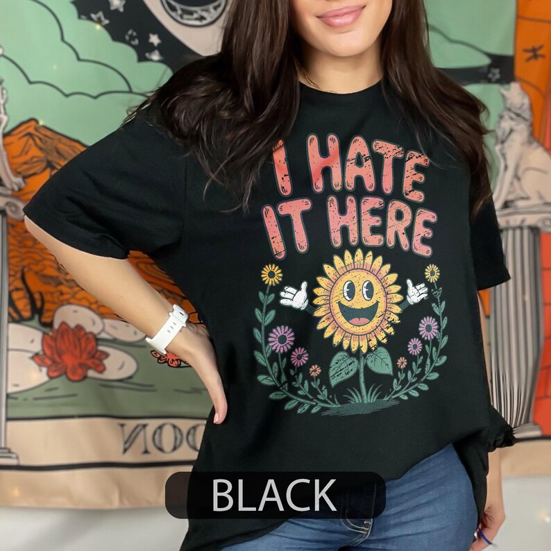 May include: Black t-shirt featuring the phrase "I HATE IT HERE" in pink and orange. A smiling sunflower graphic is below the text, surrounded by flowers and leaves. The word "BLACK" is displayed in a black rectangle at the bottom of the shirt.
