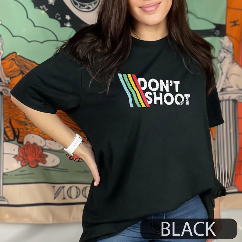 May include: Black t featuring the text "DON'T SHOOT" in white, with a colourful striped design. The word "BLACK" is at the bottom of the shirt. The shirt is a casual style.