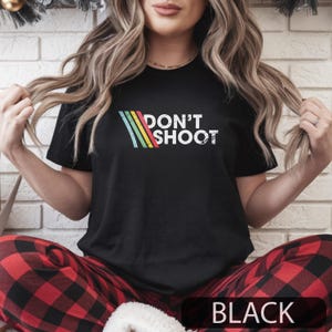 May include: Black t with the text "DON'T SHOOT" in white, with a colourful striped design. The shirt is paired with red and black plaid pyjama bottoms. The word "BLACK" is at the bottom of the image.