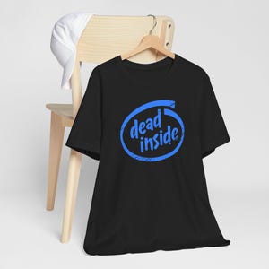 Puede incluir: Camiseta negra con un gráfico azul que dice "dead inside" en una fuente estilizada. El diseño está dentro de una forma ovalada azul. Una gorra de béisbol blanca cuelga de una silla de madera en el fondo.