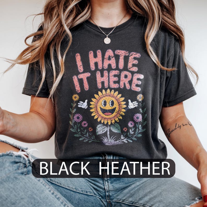 May include: Black heather t-shirt featuring the pink text "I HATE IT HERE" above a smiling sunflower graphic. The sunflower has a yellow face and petals, with white gloved hands. The text "BLACK HEATHER" is at the bottom.