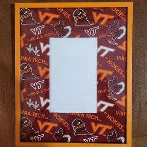 May include: Orange framed picture with a white mat. The background features a maroon pattern with the words "Virginia Tech" and "Hokies" along with the VT logo and a cartoon turkey. A decorative item for a fan.