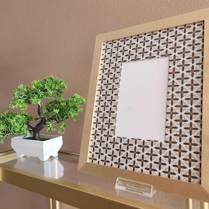 May include: A gold picture frame with a white mat and a geometric pattern on the front. The frame is sitting on a glass shelf.