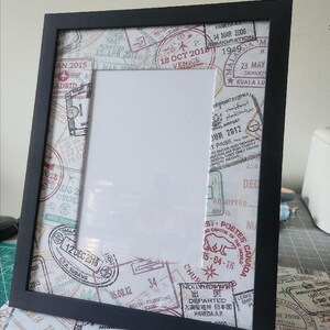 May include: A black picture frame featuring a mat board decorated with a collage of international travel passport stamps in red, blue, and black ink. The stamps include various dates, airport names, and locations like Venice, Madrid, and Haneda.