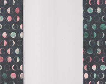 May include: A blank white notebook rests on a dark background patterned with repeating moon phase illustrations in pink, teal, and white. The notebook is centered, with the patterned background visible around its edges.