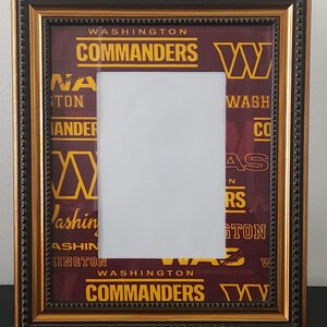 May include: A rectangular picture frame with a dark bronze and gold beaded border. The matting features a repeating pattern of the Washington Commanders logo, the team name in yellow text, and the word Washington in a burgundy and gold color scheme.