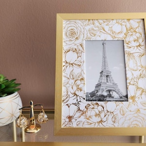 Puede incluir: Un cuadro con marco dorado que presenta una imagen en blanco y negro de la Torre Eiffel. El marco tiene un patrón floral en dorado y blanco. A la izquierda, una pequeña planta suculenta en una maceta blanca y dorada con diseño geométrico.