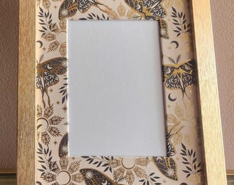 May include: A rectangular gold-colored picture frame. The frame's interior border features a cream-colored design with gold suns, moons, moths, and foliage. The frame holds a blank white space for a photo or artwork.