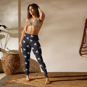 Smiling woman with light brown skin standing in a sunlit room wearing full length yoga leggings with a decorative lotus flower print in dark gray, dark purple, light blue, and light green.
