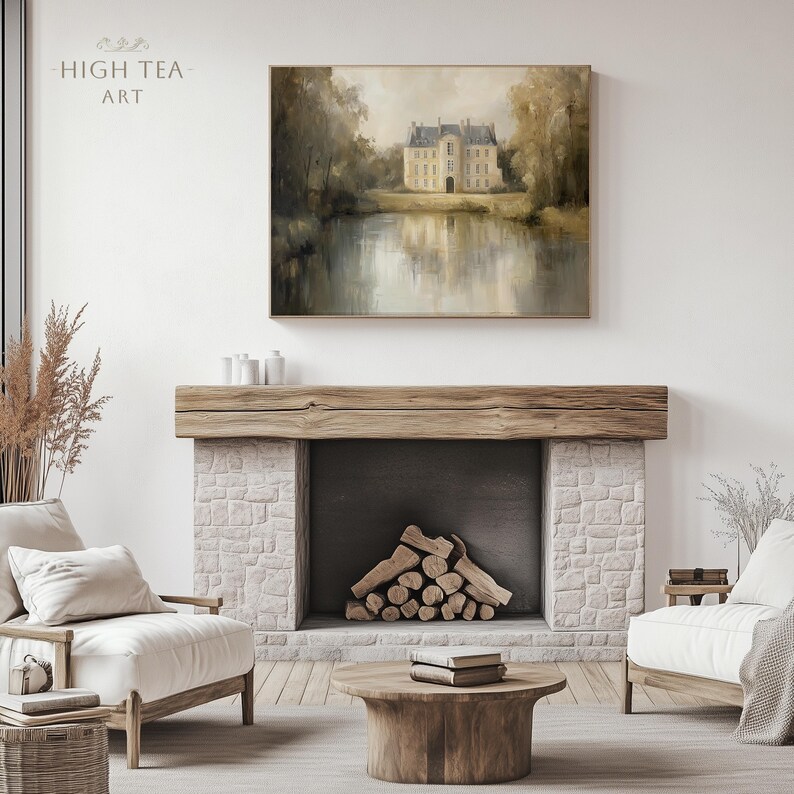 May include: A painting of a large, light-colored building with a reflection in the water in front of it. The building has a pointed roof and many windows. The painting is hanging above a fireplace with a wooden mantel and a stone surround. There are two white armchairs in front of the fireplace.