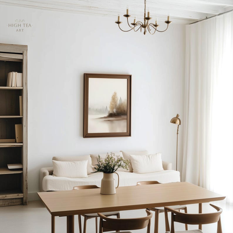 Op de afbeelding: Een lichte woonkamer met een neutrale kleurenpalet. Een ingelijste landschapsschilderij hangt boven een beige bank met witte kussens. Een houten eettafel en stoelen staan op de voorgrond. Een kroonluchter en een vloerlamp dragen bij aan de sfeer.