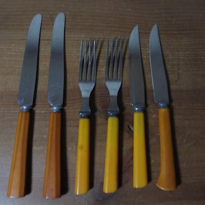 May include: A set of six vintage cutlery pieces, including knives and forks. The knives have stainless steel blades and brown handles. The forks have stainless steel tines and yellow handles. The set is arranged on a wooden surface.