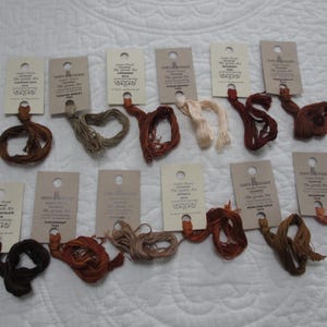 May include: A collection of embroidery floss in various shades of brown, beige, and cream, each attached to a small, rectangular tag. The tags have a hole at the top and feature printed text. The floss is arranged on a white, quilted surface.