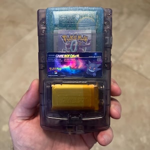 May include: A purple Game Boy Color handheld gaming console with a clear blue shell and a gold battery cover. The console has a Pokemon sticker on the front with a holographic background and a picture of a Pokemon character.