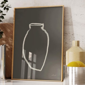 May include: A framed black and white illustration of a vase on a white background. The vase is outlined in white and has a window-like shape in the center. The frame is gold and the illustration is on a white countertop with a silver pitcher and a silver bowl with lemons.