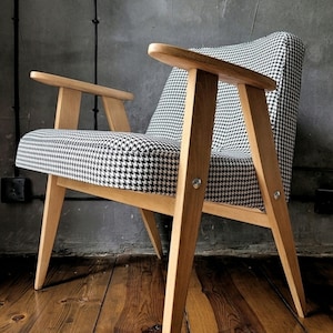 Può includere: Poltrona moderna di metà secolo con struttura in legno chiaro e rivestimento a motivo pied-de-poule. La sedia ha gambe e braccioli angolati. Lo sfondo è un muro grigio e un pavimento in legno.