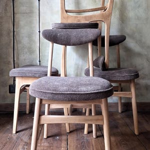 Può includere: Una collezione di sedie in legno con sedute imbottite marroni. Le sedie hanno telai e gambe in legno di colore chiaro. Le sedute sono di un marrone tenue. Le sedie sono disposte in una pila, mostrando il loro design.
