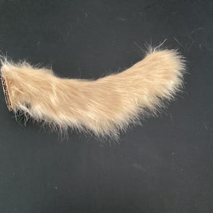 May include: A curved, light tan faux fur tail accessory featuring a dense, long-pile texture. The base of the tail is finished with a small, circular cardboard attachment point, providing a structured anchor for the soft, fluffy synthetic fibers.