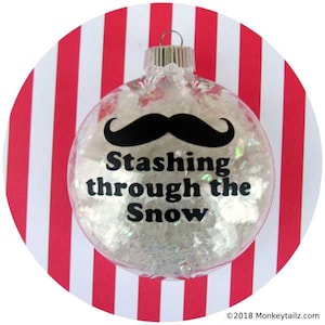 May include: A clear glass Christmas ornament with a black mustache and the text "Stashing through the Snow". The ornament is filled with white glitter and is sitting on a red and white striped background.