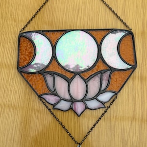 Può includere: Suncatcher in vetro colorato con un design a tripla luna sopra un fiore di loto. Ha un bordo color rame e un ciondolo di cristallo trasparente a forma di goccia iridescente. Il suncatcher è appeso a una catena.