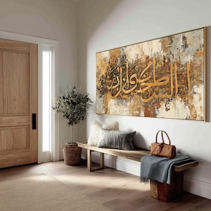 Könnte beinhalten: Große rechteckige Wandkunst mit goldener arabischer Kalligraphie auf einem strukturierten Hintergrund in Braun-, Beige- und Grautönen. Das goldgerahmte Kunstwerk hängt über einer Holzbank mit Kissen und einer Lederhandtasche.