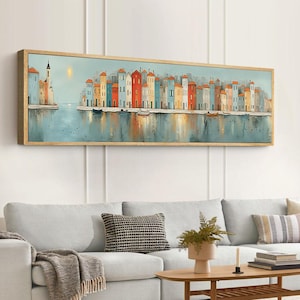 Può includere: Una stampa artistica incorniciata raffigurante una colorata scena sul lungomare. L'opera presenta una fila di edifici di vari colori, riflessi nell'acqua. Il cielo è azzurro con un sole giallo. La cornice è in legno chiaro.