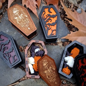May include: Wooden coffin-shaped boxes in various colors, some with carved designs and filled with Halloween-themed items like pumpkins and ghosts. One box has the text "NEVER BETTER" and a skeleton design.