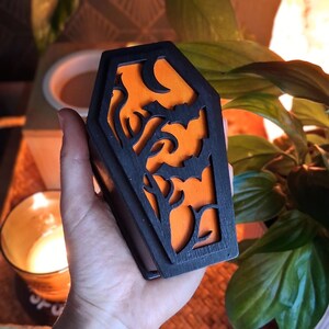 May include: A black coffin-shaped box with an orange interior, featuring cut-out designs of bats, trees, and a crescent moon. The box is held in a hand, with a candle and plants in the background. The design is perfect for Halloween.