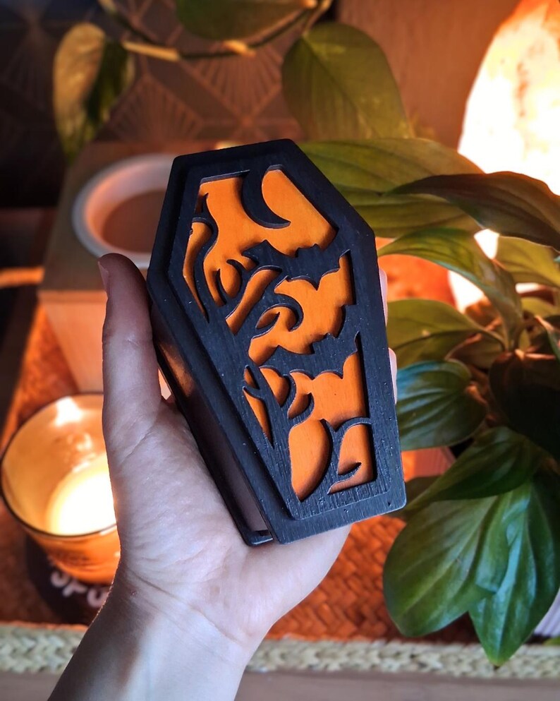 May include: A black coffin-shaped box with an orange interior, featuring cut-out designs of bats, trees, and a crescent moon. The box is held in a hand, with a candle and plants in the background. The design is perfect for Halloween.