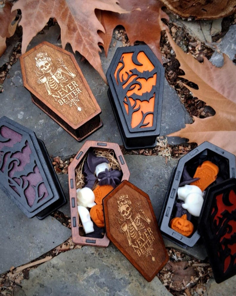 May include: Wooden coffin-shaped boxes in various colors, some with carved designs and filled with Halloween-themed items like pumpkins and ghosts. One box has the text "NEVER BETTER" and a skeleton design.