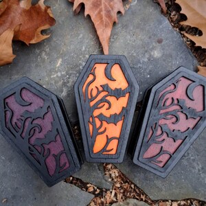 May include: Three wooden coffin-shaped boxes with black frames and carved designs. The center box has an orange interior with a bat and tree design. The other two have purple and red interiors with similar designs. The boxes are on a stone surface with leaves.