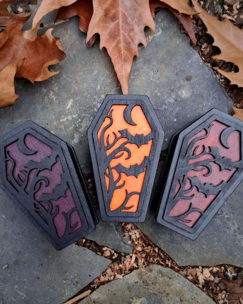 May include: Three wooden coffin-shaped boxes with black frames and carved designs. The center box has an orange interior with a bat and tree design. The other two have purple and red interiors with similar designs. The boxes are on a stone surface with leaves.