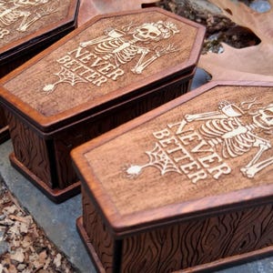 May include: Wooden coffin-shaped boxes with a dark brown finish. Each box features a carved skeleton design on the lid, along with the words "NEVER BETTER" and a spiderweb. The boxes have a wood grain texture.
