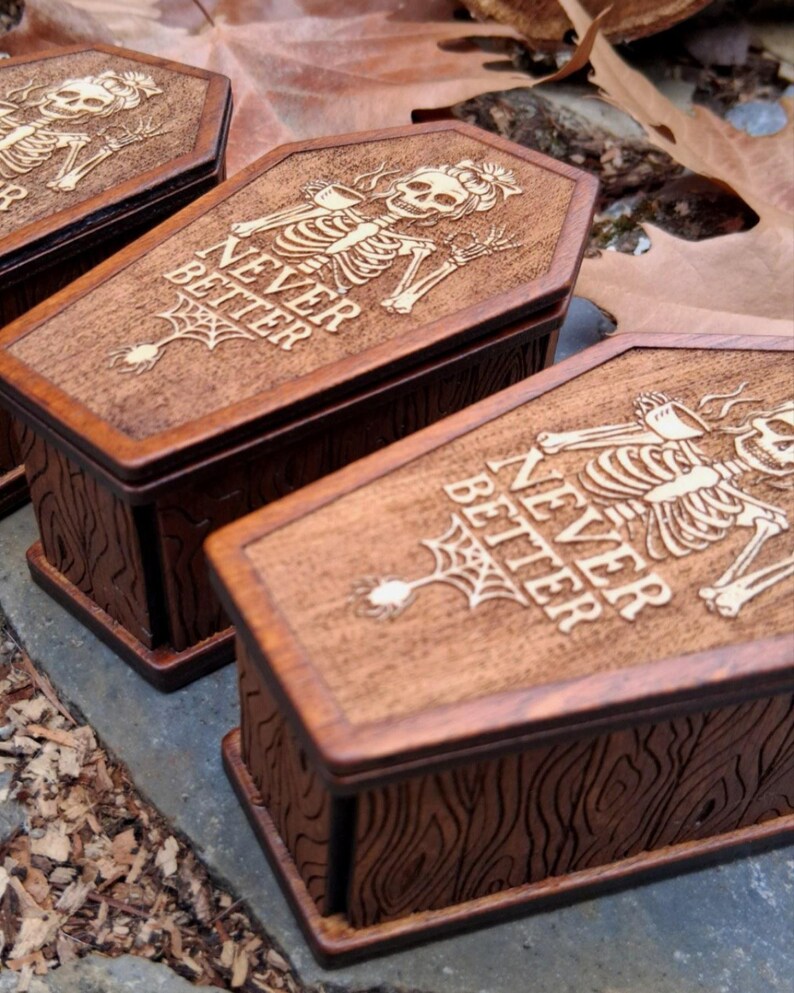 May include: Wooden coffin-shaped boxes with a dark brown finish. Each box features a carved skeleton design on the lid, along with the words "NEVER BETTER" and a spiderweb. The boxes have a wood grain texture.