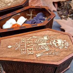 May include: Wooden coffin-shaped boxes with engraved skeleton and spiderweb designs. One box is open, revealing colorful contents. The text on the lid reads "Never Better". The boxes are a dark brown color.