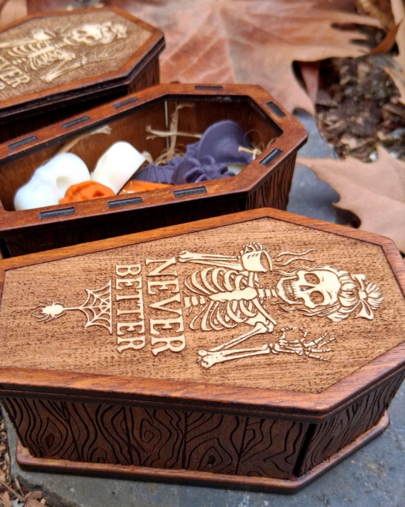 May include: Wooden coffin-shaped boxes with engraved skeleton and spiderweb designs. One box is open, revealing colorful contents. The text on the lid reads "Never Better". The boxes are a dark brown color.