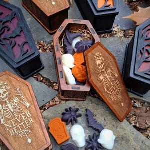 May include: Wooden coffin-shaped boxes with carved designs and the text "NEVER BETTER". The boxes are open, revealing Halloween-themed soaps in the shapes of ghosts, spiders, and pumpkins. The boxes are brown, black, and orange.