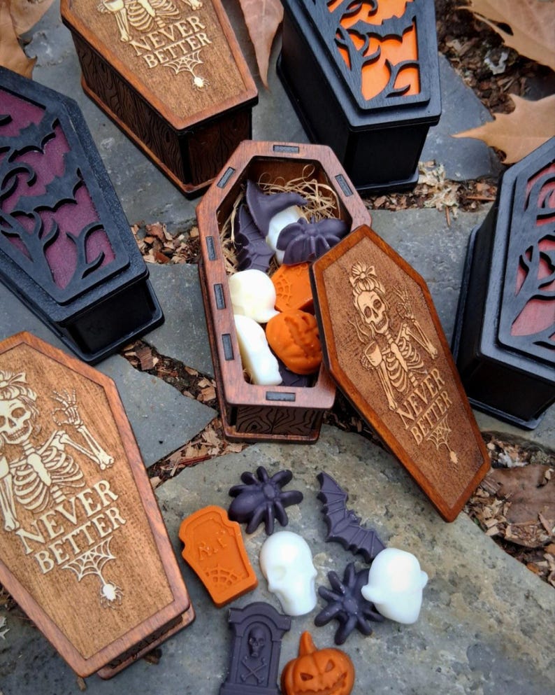 May include: Wooden coffin-shaped boxes with carved designs and the text "NEVER BETTER". The boxes are open, revealing Halloween-themed soaps in the shapes of ghosts, spiders, and pumpkins. The boxes are brown, black, and orange.
