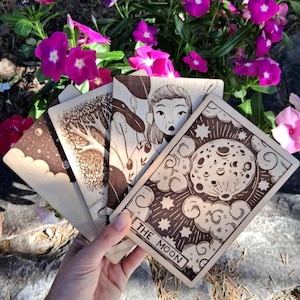 Puede incluir: Una mano sostiene una baraja de cartas de tarot de madera con intrincados diseños pirograbados. Las cartas presentan ilustraciones de un árbol, una mujer y la luna, con el texto "THE MOON". El fondo incluye vibrantes flores rosas y vegetación.