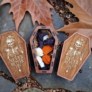 May include: Three wooden coffin-shaped boxes. Two boxes are closed, featuring a skeleton with a teacup and the text "NEVER BETTER". The open box contains Halloween-themed items, including a witch hat, spider, and pumpkin.
