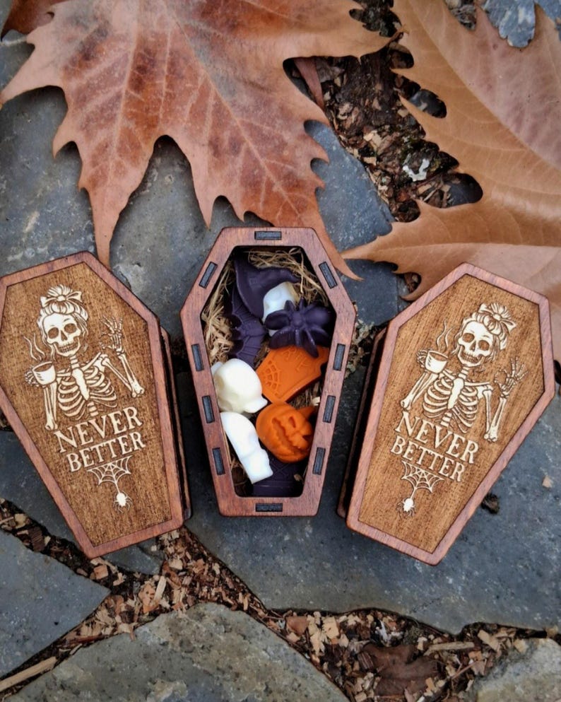 May include: Three wooden coffin-shaped boxes. Two boxes are closed, featuring a skeleton with a teacup and the text "NEVER BETTER". The open box contains Halloween-themed items, including a witch hat, spider, and pumpkin.