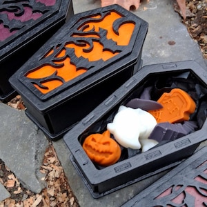 May include: Black coffin-shaped boxes with carved designs and orange, purple, and black interiors. One box is open, revealing Halloween-themed soaps, including a pumpkin, ghost, and bat. The boxes are displayed on a stone surface.